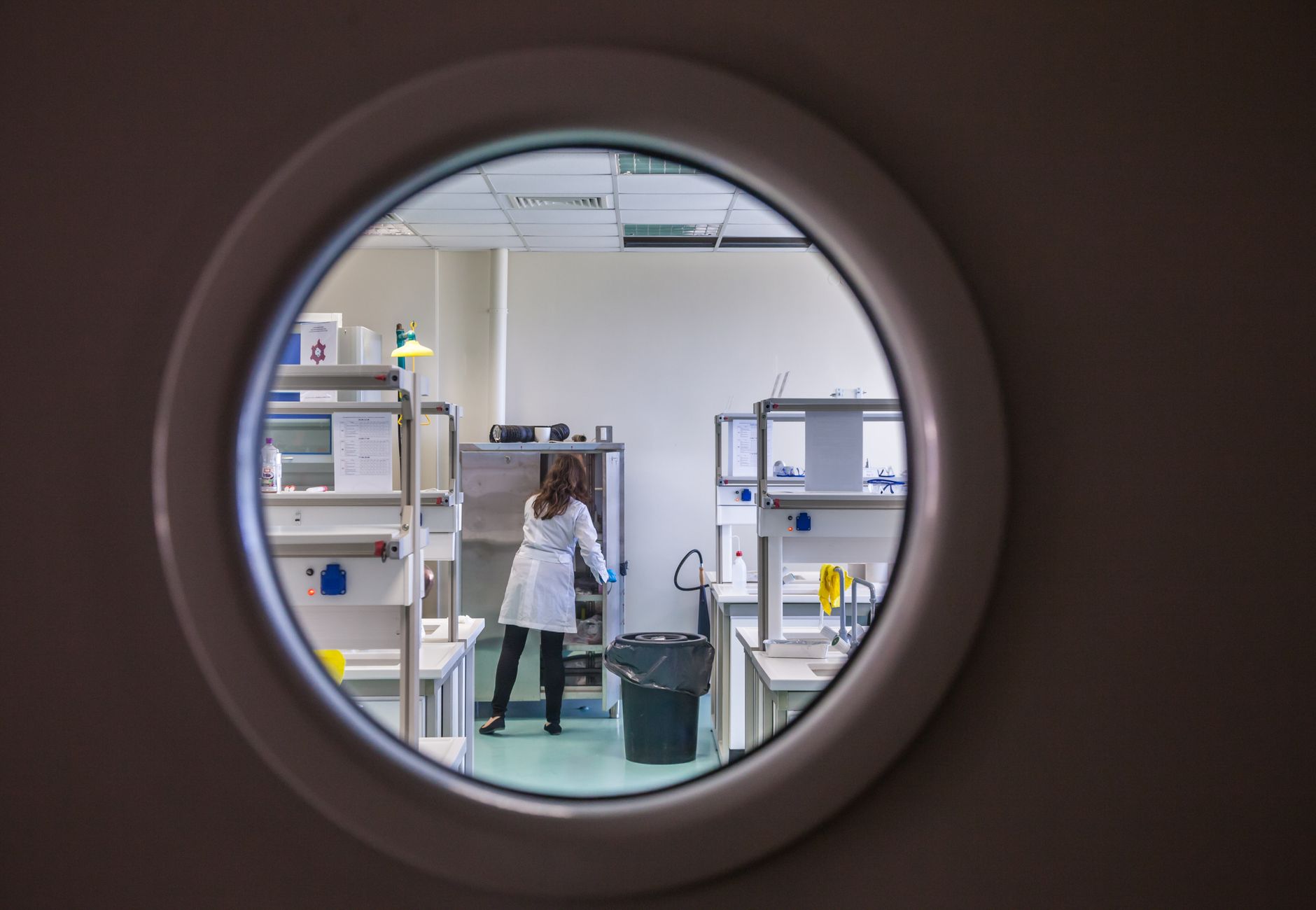 A female scientist in a lab coat working inside a state-of-the-art laboratory through a circular window view.