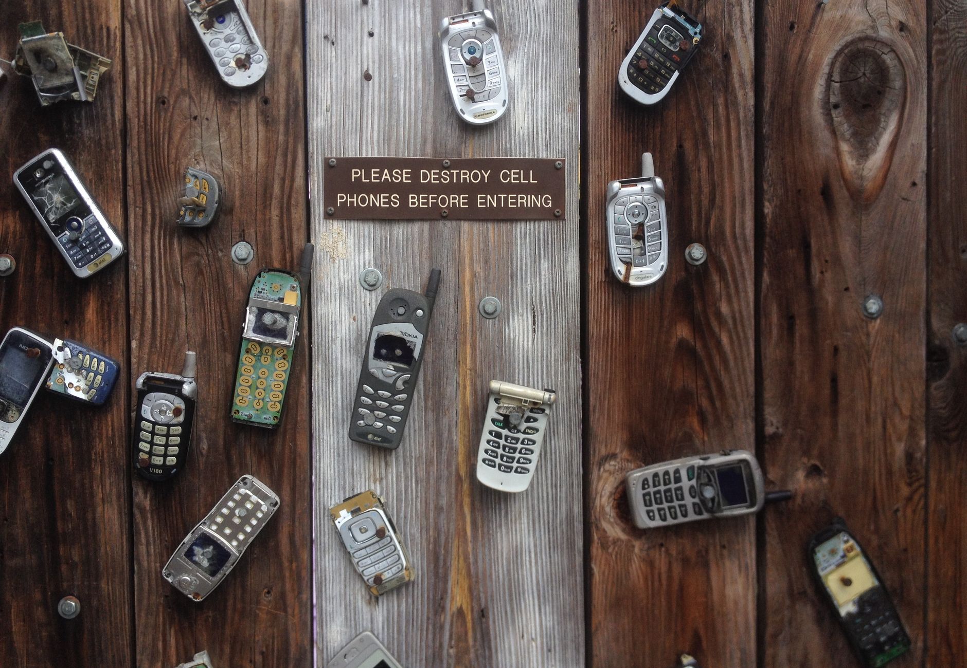 Collection of vintage cell phones attached to a rustic wooden wall with a humorous sign.